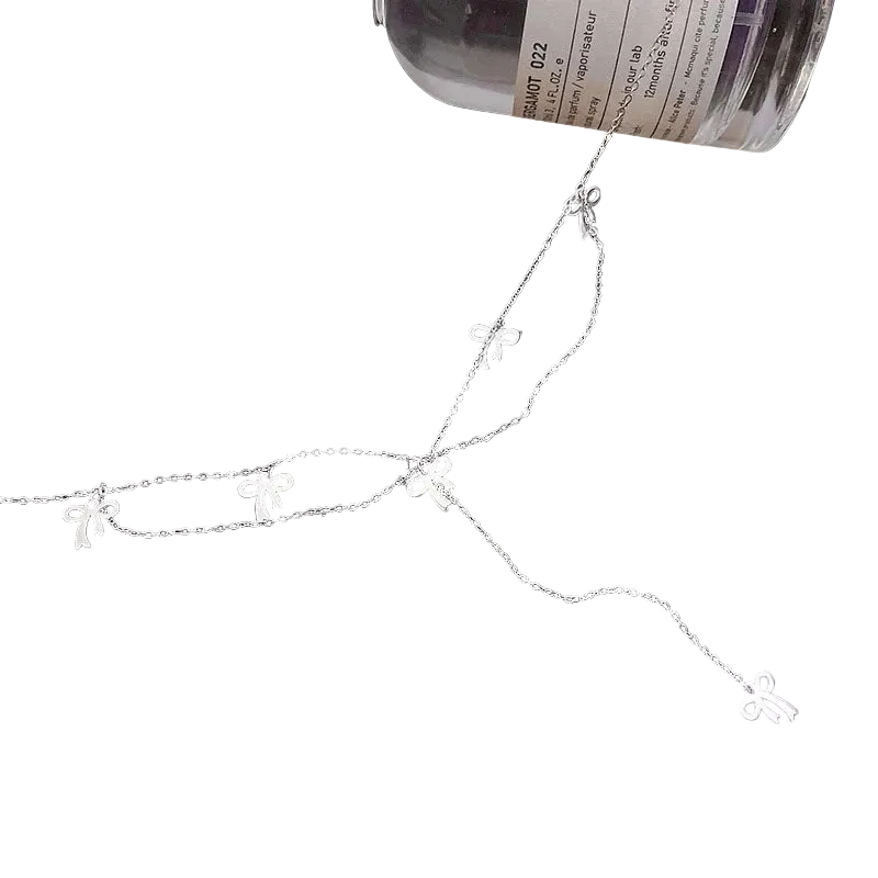A delicate silver chain necklace featuring five small bow-shaped charms, displayed on a black background with a bottle partially visible at the top right.