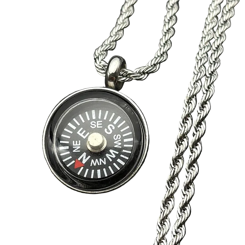 A silver compass pendant necklace with a detailed compass face, resting on a dark textured surface next to a silver chain.