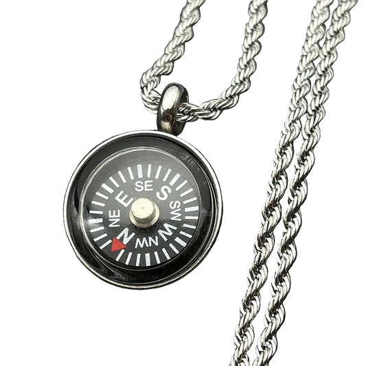 A silver compass pendant necklace with a detailed compass face, resting on a dark textured surface next to a silver chain.