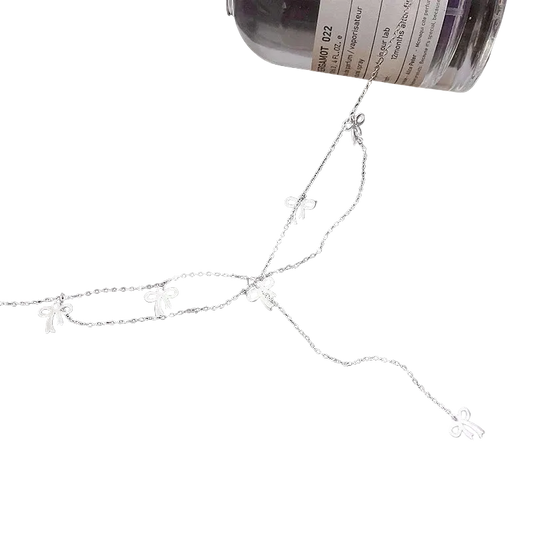 A delicate silver chain necklace featuring five small bow-shaped charms, displayed on a black background with a bottle partially visible at the top right.