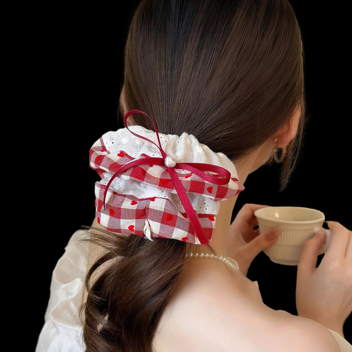 A woman shown from behind wearing a red and white gingham hair scrunchie with a matching red ribbon bow, holding a small cup in a softly lit indoor setting.