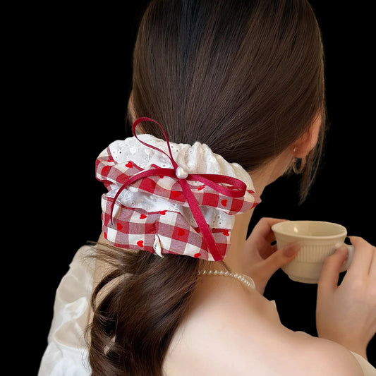 A woman shown from behind wearing a red and white gingham hair scrunchie with a matching red ribbon bow, holding a small cup in a softly lit indoor setting.