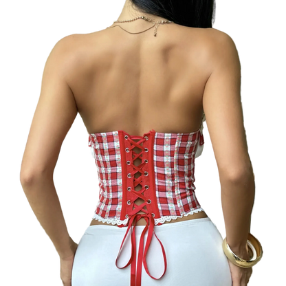A woman shown from the back wearing a red and white gingham strapless corset top with lace-up back detailing and white bottoms, standing indoors near a patio door and chair.