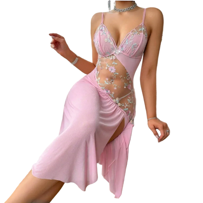 A woman models a light pink satin slip dress with floral embroidery and sheer mesh panels, sitting on a white textured armchair in a bright room.