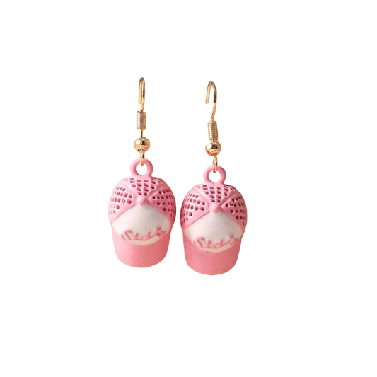A pair of dangling earrings displayed on a beige jewelry bust. Each earring has a gold fishhook wire and a pink charm shaped like a cupcake, with a pink lace-knit top and a white cake body featuring pink frosting details.