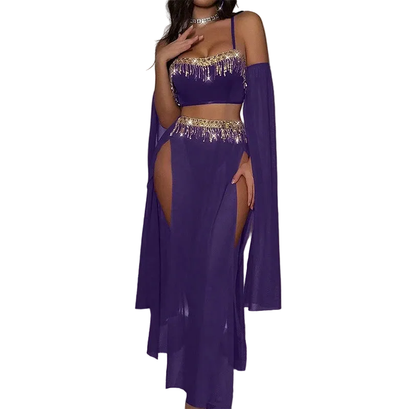 A woman models a purple belly dance costume set featuring a crop top with gold fringe, a matching high-slit skirt, and a sheer long cape, in a room with a pink neon light.