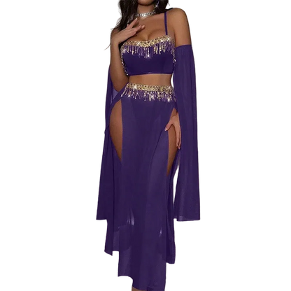 A woman models a purple belly dance costume set featuring a crop top with gold fringe, a matching high-slit skirt, and a sheer long cape, in a room with a pink neon light.