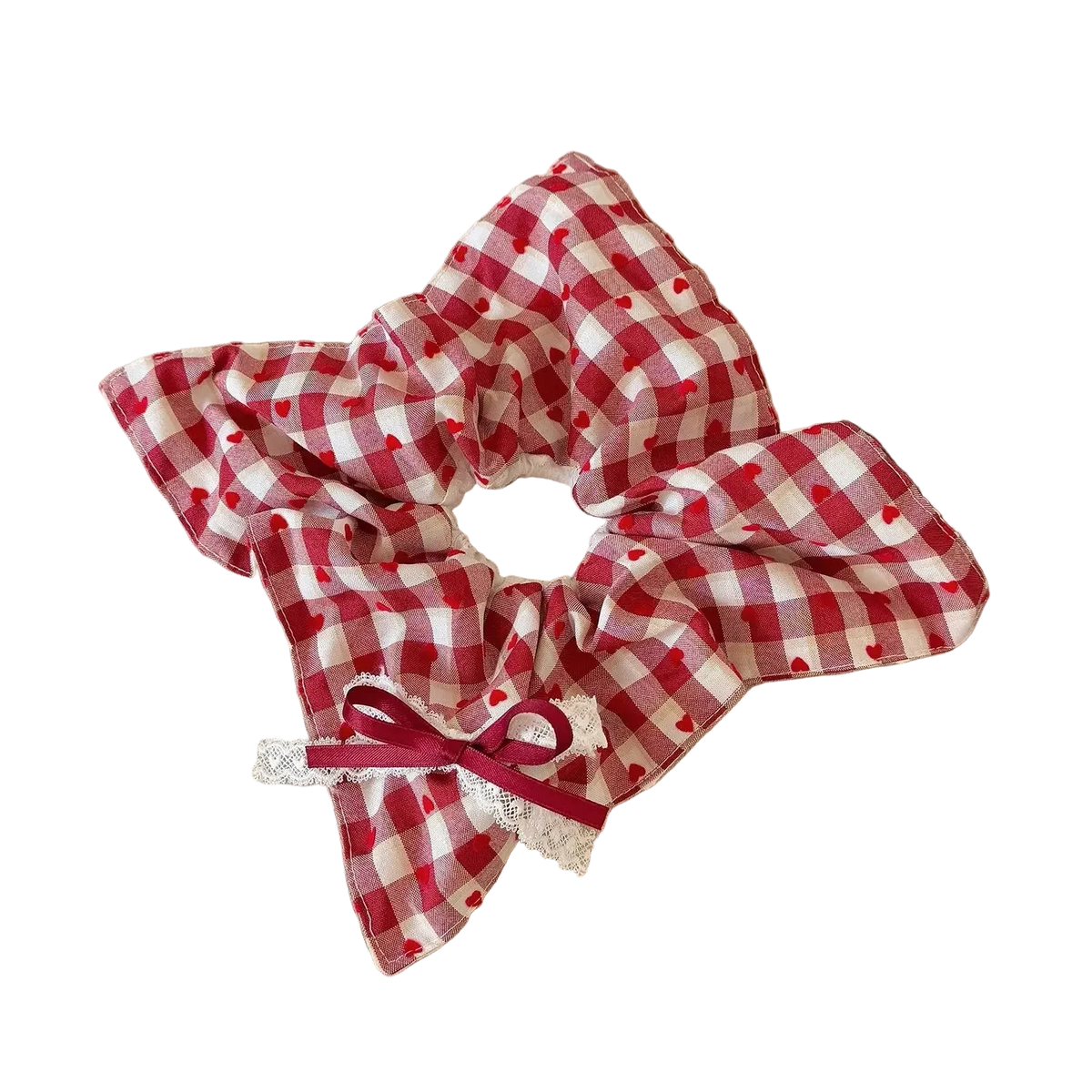 A red and white gingham fabric hair scrunchie with small red hearts and a white lace trim, tied with a red ribbon bow, displayed on a beige book on a wooden table.