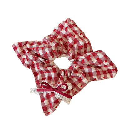A red and white gingham fabric hair scrunchie with small red hearts and a white lace trim, tied with a red ribbon bow, displayed on a beige book on a wooden table.