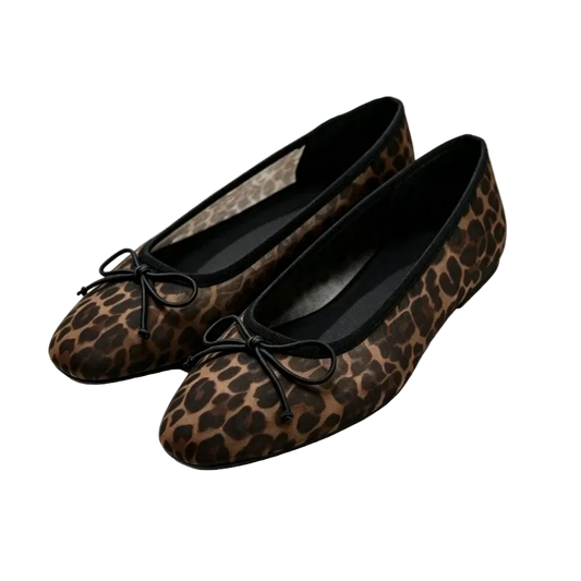 A pair of women's flat ballet flats in a brown and black leopard print, featuring black fabric bows on the toes, displayed on a white platform.
