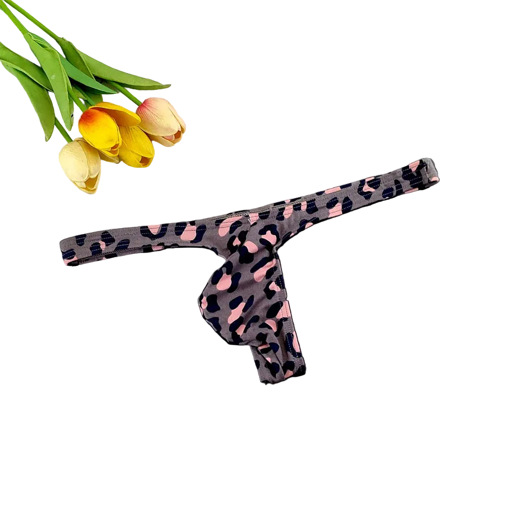 A single thong-style underwear piece with a dark gray base and pink and black leopard print, laid flat on a dark surface with tulips and a white grid tile as styling props.