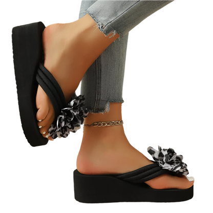 Black wedge sandals with floral details worn by a person on a wooden floor.