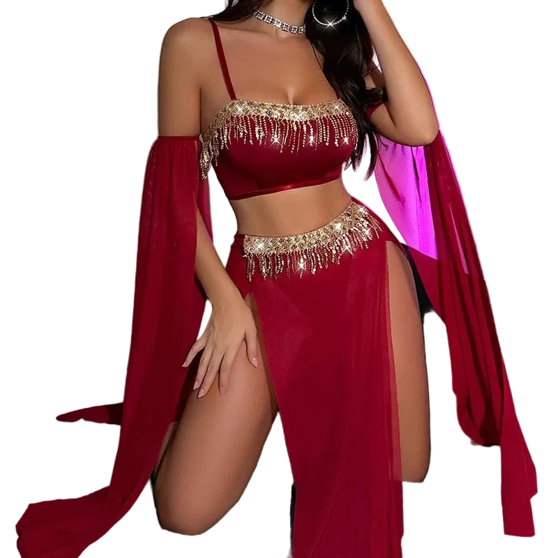 A woman models a deep red belly dance costume set featuring a crop top with gold fringe, a matching high-slit skirt, and a sheer draped cape, posed on a black couch against a wall with pink ambient lighting.