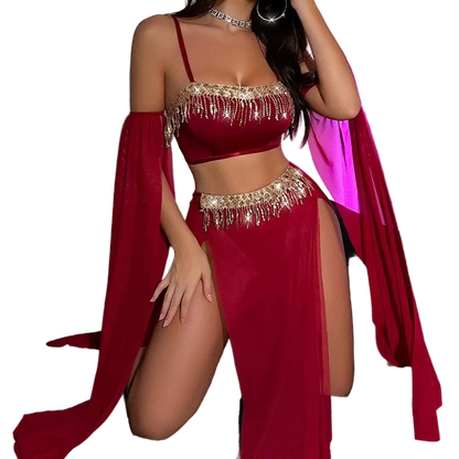 A woman models a deep red belly dance costume set featuring a crop top with gold fringe, a matching high-slit skirt, and a sheer draped cape, posed on a black couch against a wall with pink ambient lighting.