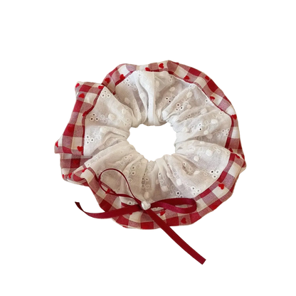 A fabric hair scrunchie with a red and white gingham border, white eyelet lace center, and a red ribbon bow, resting on a beige book on a wooden table.