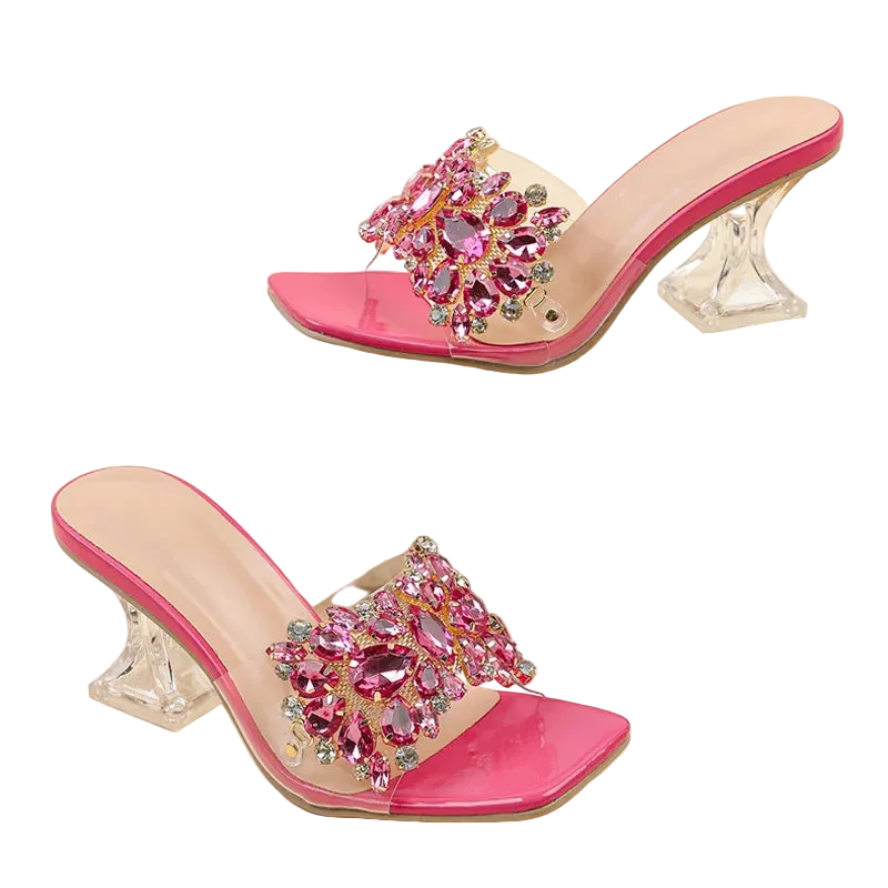 A pair of women's heeled mule sandals with bright pink patent leather trim, clear transparent straps, and large pink crystal embellishments on the toes, displayed on a beige background with one shoe resting on a white block.