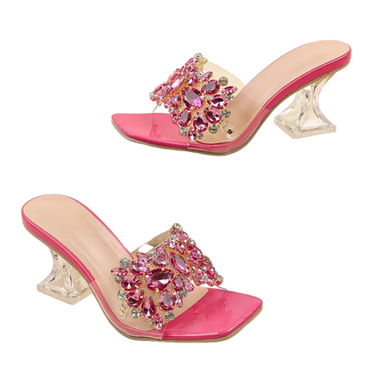 A pair of women's heeled mule sandals with bright pink patent leather trim, clear transparent straps, and large pink crystal embellishments on the toes, displayed on a beige background with one shoe resting on a white block.