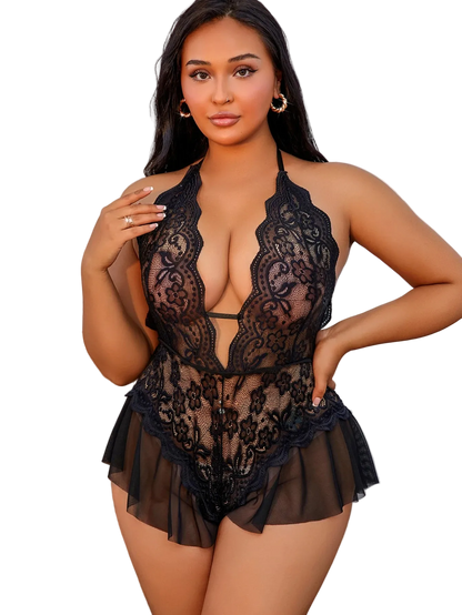 A woman modeling a black lace and sheer mesh halter bodysuit with ruffled shorts in an indoor setting with a white paneled wall and a wooden cabinet.