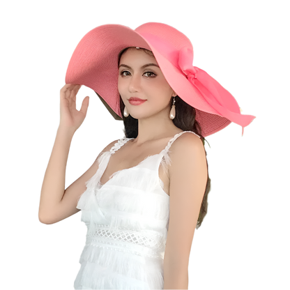 Woman wearing a large pink hat and white dress outdoors with greenery.