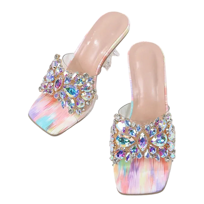 A pair of women's heeled mule sandals with iridescent pastel rainbow footbeds and large crystal embellishments on the straps, displayed on a white marble surface with a denim bag and woven strap as props.