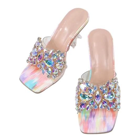A pair of women's heeled mule sandals with iridescent pastel rainbow footbeds and large crystal embellishments on the straps, displayed on a white marble surface with a denim bag and woven strap as props.