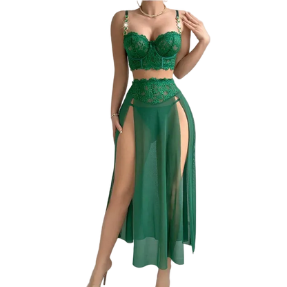 A woman models a two-piece green lace and sheer maxi dress set against a solid gray background. The set includes a green lace bralette with gold chain straps and a matching high-waisted sheer skirt with a thigh-high slit.