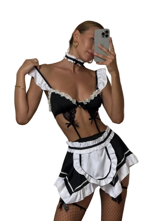 A woman takes a mirror selfie wearing a black and white maid costume set with ruffles, a lace bra top, a mini skirt, and a matching choker. She is holding a silver smartphone in a bedroom setting.