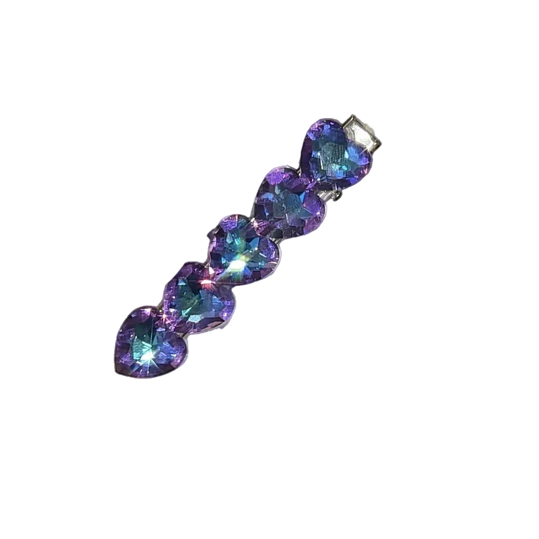 A heart-shaped hair claw clip adorned with four iridescent purple and blue rhinestones, attached to a white display card held by a hand with a glittery manicure.