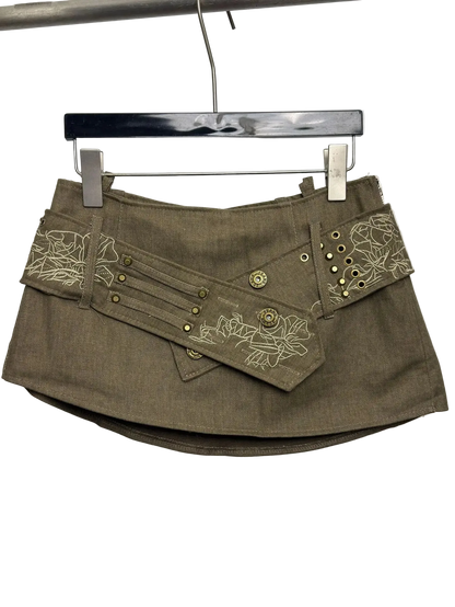An olive brown mini skirt with decorative floral embroidery and brass grommets, hanging on a black hanger against a plain white background.
