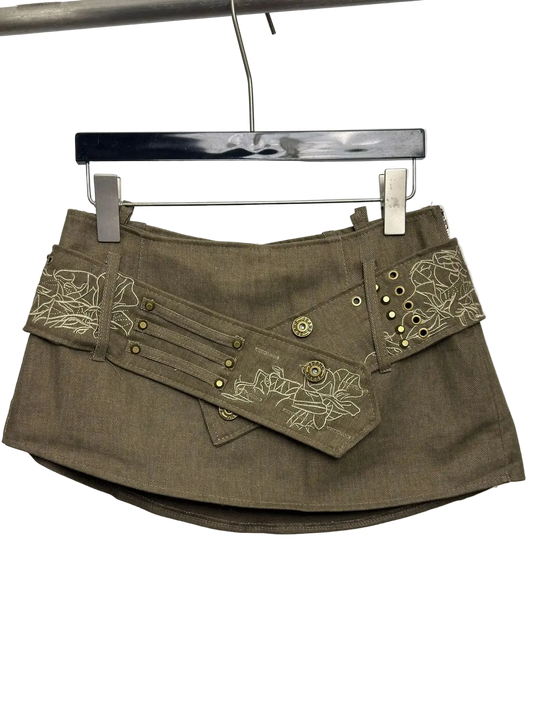 An olive brown mini skirt with decorative floral embroidery and brass grommets, hanging on a black hanger against a plain white background.