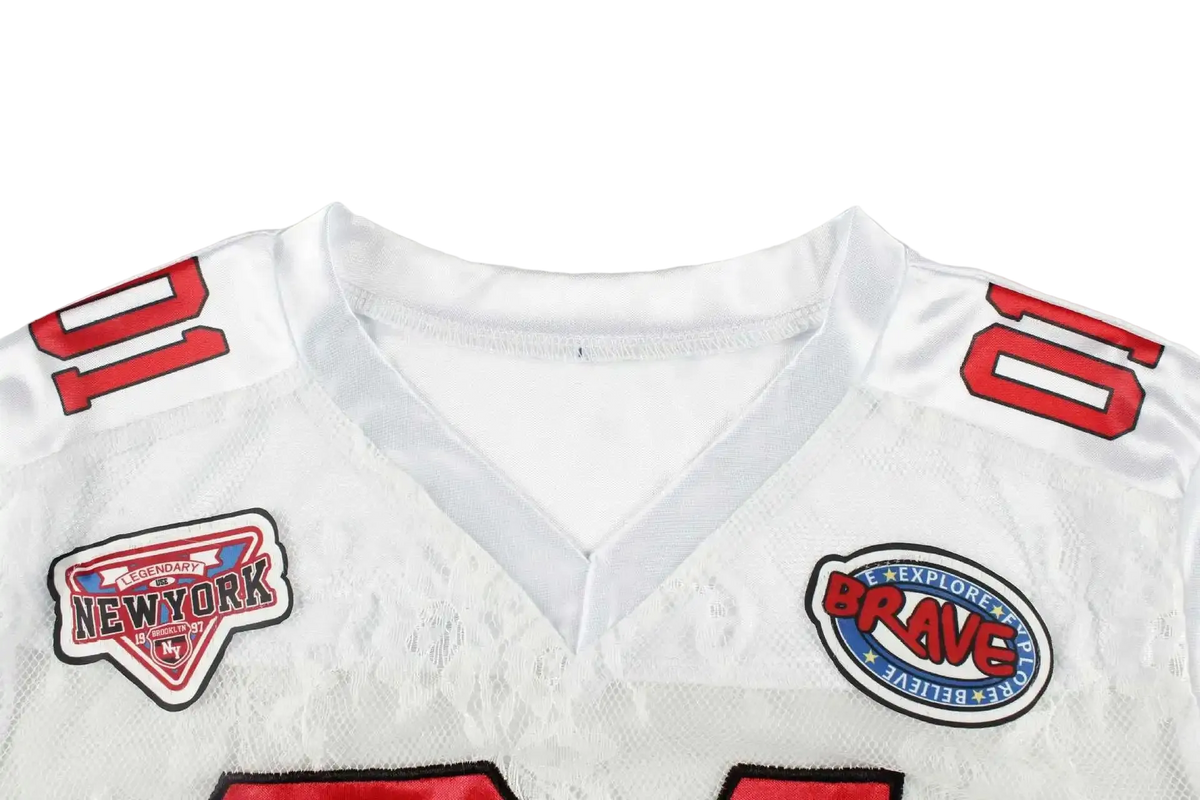 Close-up of a white sports jersey with red shoulder numbers and two embroidered patches on the chest, photographed against a light gray background.