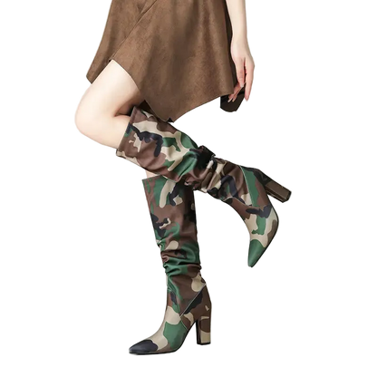A model's legs wearing a pair of knee-high camouflage boots with a pointed toe and block heel, paired with a brown dress against a clean studio background.