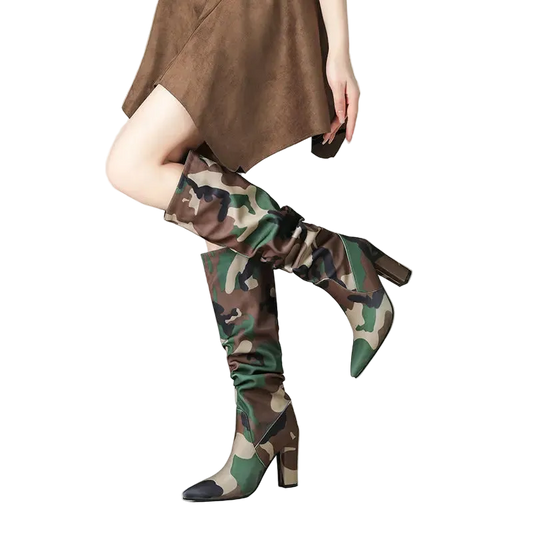 A model's legs wearing a pair of knee-high camouflage boots with a pointed toe and block heel, paired with a brown dress against a clean studio background.