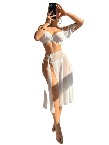 A woman takes a mirror selfie wearing a white two-piece lingerie set consisting of a bra and a sheer high-slit skirt, in a brightly lit room with strong sunlight and shadows.
