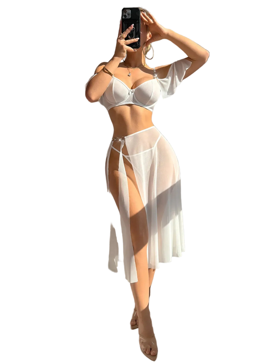 A woman takes a mirror selfie wearing a white two-piece lingerie set consisting of a bra and a sheer high-slit skirt, in a brightly lit room with strong sunlight and shadows.