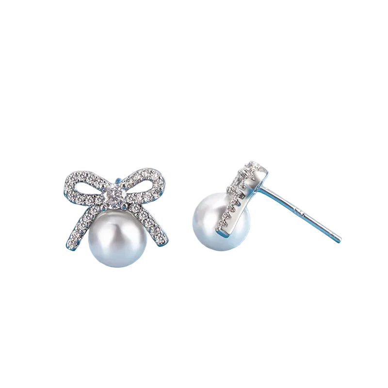 A pair of silver stud earrings featuring a bow-shaped setting paved with clear crystals and a white pearl hanging below, displayed on a light blue surface.
