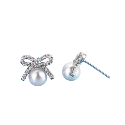 A pair of silver stud earrings featuring a bow-shaped setting paved with clear crystals and a white pearl hanging below, displayed on a light blue surface.
