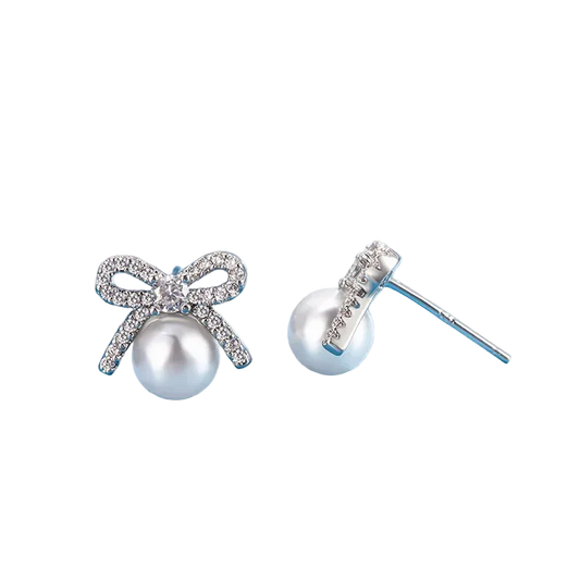 A pair of silver stud earrings featuring a bow-shaped setting paved with clear crystals and a white pearl hanging below, displayed on a light blue surface.
