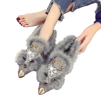 A pair of gray faux-fur slip-on flats with pointed metallic gold toes, rhinestone fox-face details, and gold star embellishments, shown being worn with denim jeans on a gray floor.