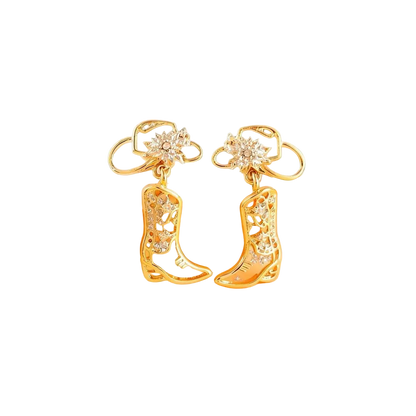 A pair of gold-toned cowboy boot earrings with clear rhinestone embellishments, displayed on a soft beige and peach background.