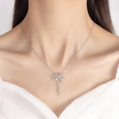 A delicate silver necklace with a bow-shaped pendant set with clear stones, worn by a model in a white top against a light gray background.