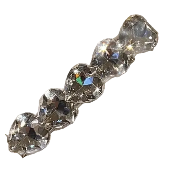 A close-up of a clear faceted heart-shaped rhinestone barrette or hair clip attached to a white display card, held by a hand with a glittery manicure in an indoor setting.