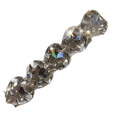 A close-up of a clear faceted heart-shaped rhinestone barrette or hair clip attached to a white display card, held by a hand with a glittery manicure in an indoor setting.