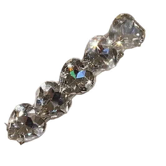 A close-up of a clear faceted heart-shaped rhinestone barrette or hair clip attached to a white display card, held by a hand with a glittery manicure in an indoor setting.
