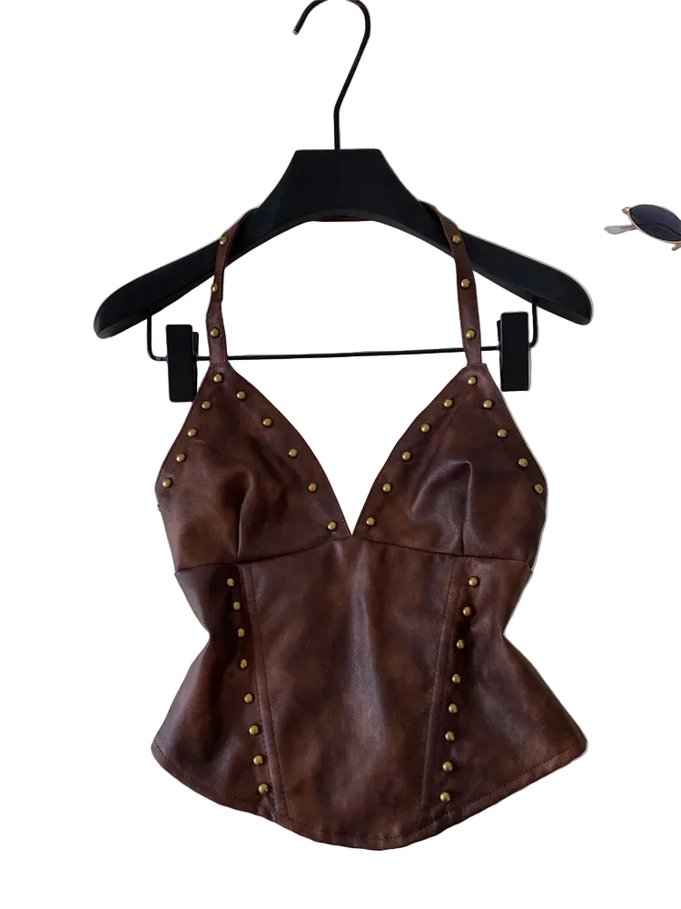 A brown faux-leather halter top with gold-tone metal studs, hanging on a black hanger against a light gray background with a laptop and sunglasses partially visible.