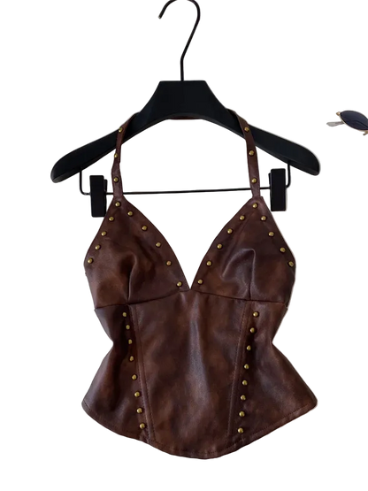 A brown faux-leather halter top with gold-tone metal studs, hanging on a black hanger against a light gray background with a laptop and sunglasses partially visible.