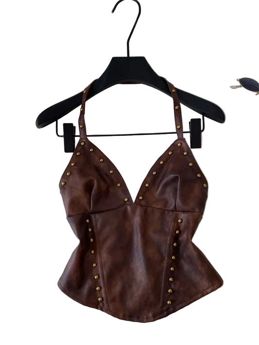 A brown faux-leather halter top with gold-tone metal studs, hanging on a black hanger against a light gray background with a laptop and sunglasses partially visible.