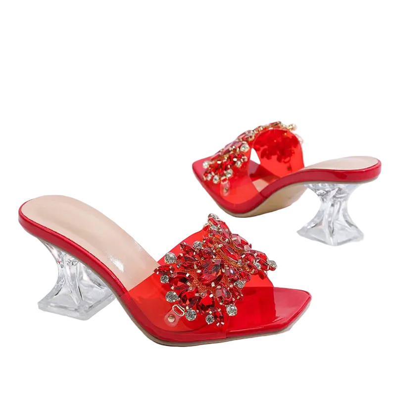 Red open-toe mule heels with crystal-embellished straps and clear acrylic block heels, shown on a white background with dried flowers in the corner.