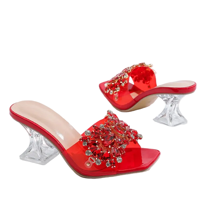 Red open-toe mule heels with crystal-embellished straps and clear acrylic block heels, shown on a white background with dried flowers in the corner.