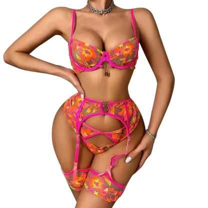 A woman modeling a bright pink and orange floral lingerie set with a matching garter belt and thigh-high stockings against a plain white background.